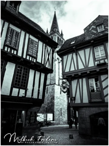 Sous les poutres du temps, Vannes murmure Dans l’écrin noir et blanc d’un matin sans hâte, Deux maisons penchées, au charme qui perdure, Veillent sur les pas lents d’une ruelle étroite. Le bois craque en silence, le pavé se souvient, Des sabots, des rires, des ombres disparues, Et là, dans l’axe noble du clocher ancien, La pierre s’élève, grave, vers la voûte inconnue. Un fer forgé s’élance, comme un bras qui salue, Peut-être un estaminet, ou l’écho d’un métier, Et les passants, flous, comme des âmes venues Cueillir un peu de paix dans ce décor figé. Vannes, tu es poème, tu es souffle et mémoire, Chaque façade est un vers, chaque ruelle un espoir. Et dans ton gris sculpté, dans ton bois qui soupire, C’est tout un vieux roman que l’on croit voir s’écrire.